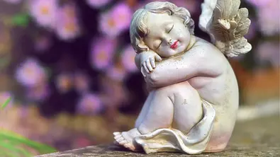 Kleine Engelstatue sitzt auf einer Steinmauer vor einem Blumenstrauch - Foto: izzzy71 / AdobeStock