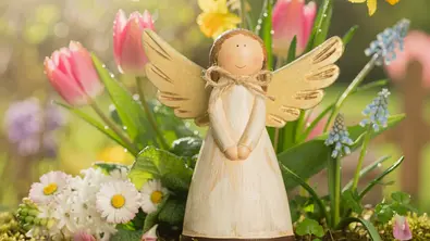 Eine kleine angemalte Engelfigur aus Holz umgeben von bunten Frühlingsblumen - Foto: AdobeFirefly (generiert mit KI)