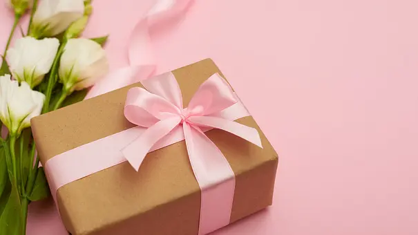 Geburtstagsgeschenk mit rosa Schleife vor einem rosa Hintergrund neben einem Strauß mit weißen Blumen - Foto: ArtFamily/ AdobeStock Geburtstagsgeschenk mit rosa Schleife vor einem rosa Hintergrund neben einem Strauß mit weißen Blumen - Foto: ArtFamily/ AdobeStock