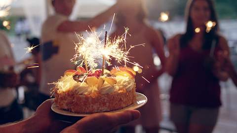 Geburtstagstorte mit Obst und Kerzen verziert wird zu einer Partygesellschaft getragen - Foto: janiecbros / iStock