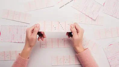 Frau hält einen Lotto-Schein in der Hand. Vor ihr liegen diverse weitere. - Foto: LIGHTFIELD STUDIOS / AdobeStock