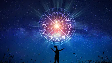 An einem Feldrand erstreckt sich ein blauer Sternenhimmel mit dem astrologischen Tierkreiszeichen in leuchtenden Farben. Davor ist die Silhouette eine tanzenden Frau zu sehen. - Foto: sarayut/iStock