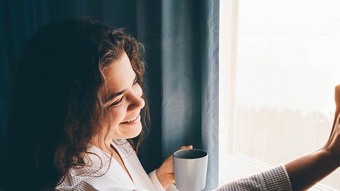 Frau schaut mit Kaffee in der Hand aus dem Fenster - Foto: Maria Korneeva/iStock