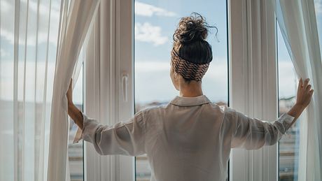 Frau öffnet die Vorhänge und blickt aus dem Fenster - Foto: iStock/Milan Markovic