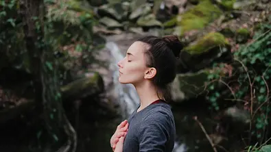 Frau, die mit den Händen auf der Brust in einem Wald durchatmet - Foto: Yolya / iStock