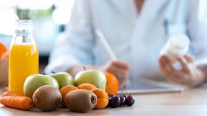 Ärztin schreibt im Hintergrund ein ärztliches Rezept vorne auf dem Schreibtisch steht eine Falsche Orangensaft umringt von Früchten. - Foto: nensuria / iStock