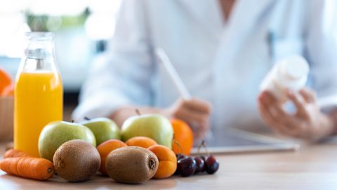 Ärztin schreibt im Hintergrund ein ärztliches Rezept vorne auf dem Schreibtisch steht eine Falsche Orangensaft umringt von Früchten. - Foto: nensuria / iStock