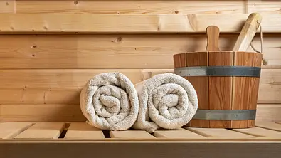Ein Holzeimer und zwei weiße Handtücher auf einer Saunabank - Foto: Alessandra Finding / iStock