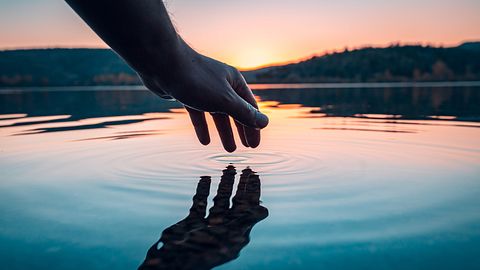 Ein See mit einem Sonnenuntergang, in dem eine Hand eingetaucht wird - Foto: iStock/ SHODOgraphy