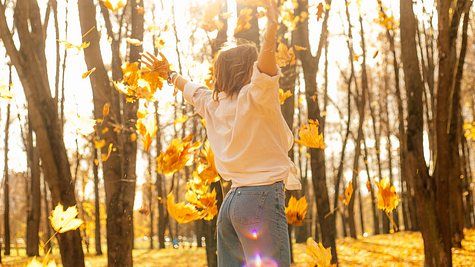 Rückansicht einer jungen Frau, die bei einem Herbstspaziergang Blätter in die Luft wirft. - Foto: Tatsiana Volkava / iStock