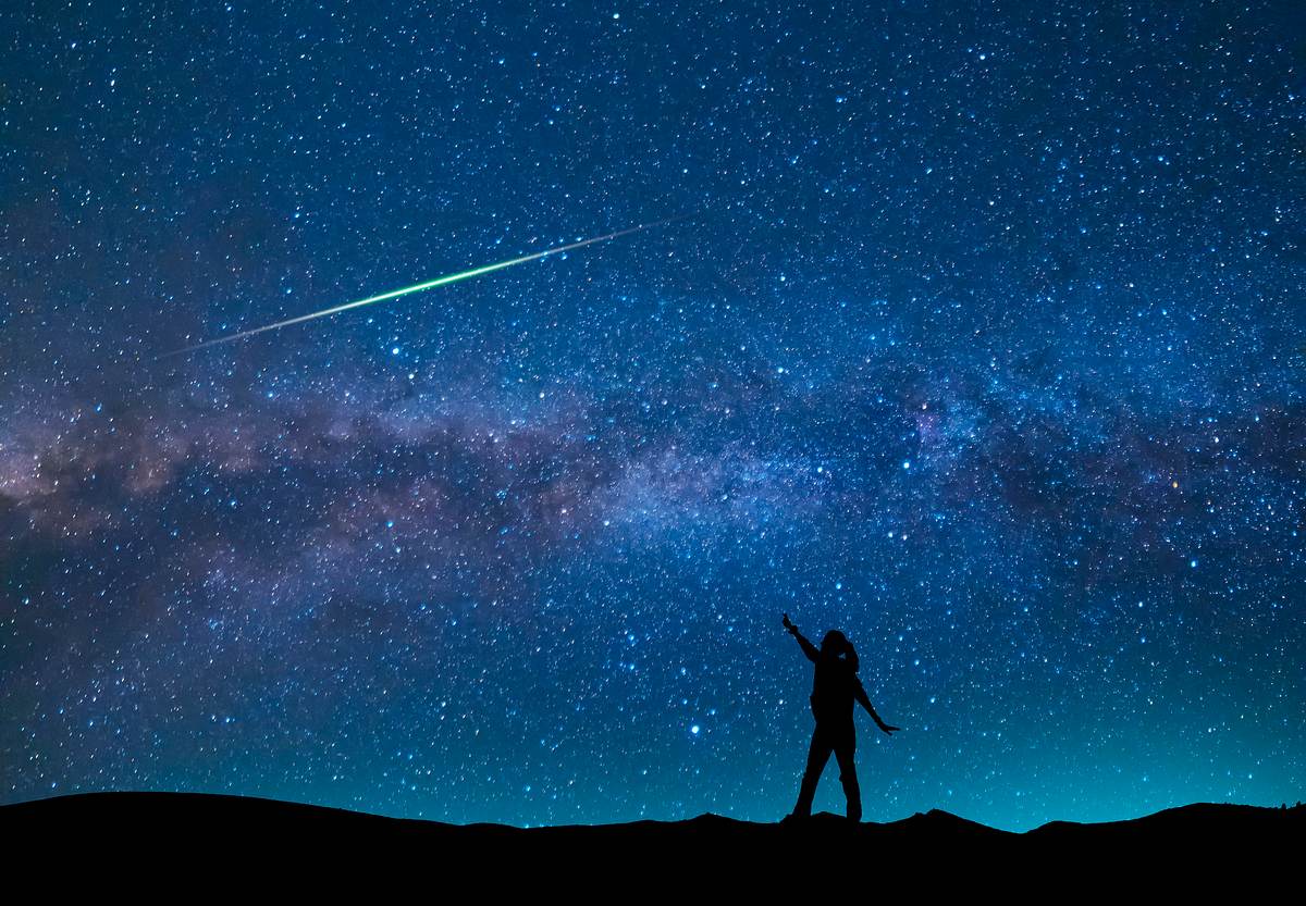 Person steht mit ausgebreiteten Armen vor einem blauen Sternenhimmel an dem eine Sternschnuppe vorüberzieht. 