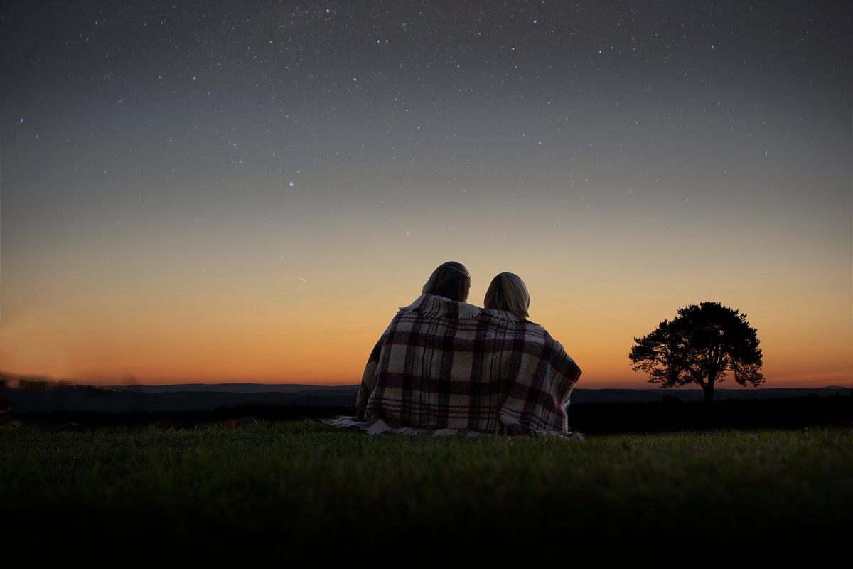 Zwei Personen sitzen auf einer Wiese mit einer Decke um den Schultern und schauen auf den Sternenhimmel.