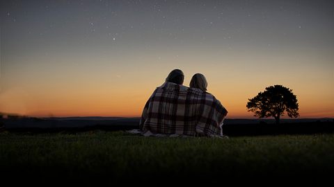 Zwei Personen sitzen auf einer Wiese mit einer Decke um den Schultern und schauen auf den Sternenhimmel. - Foto: SolStock/iStock