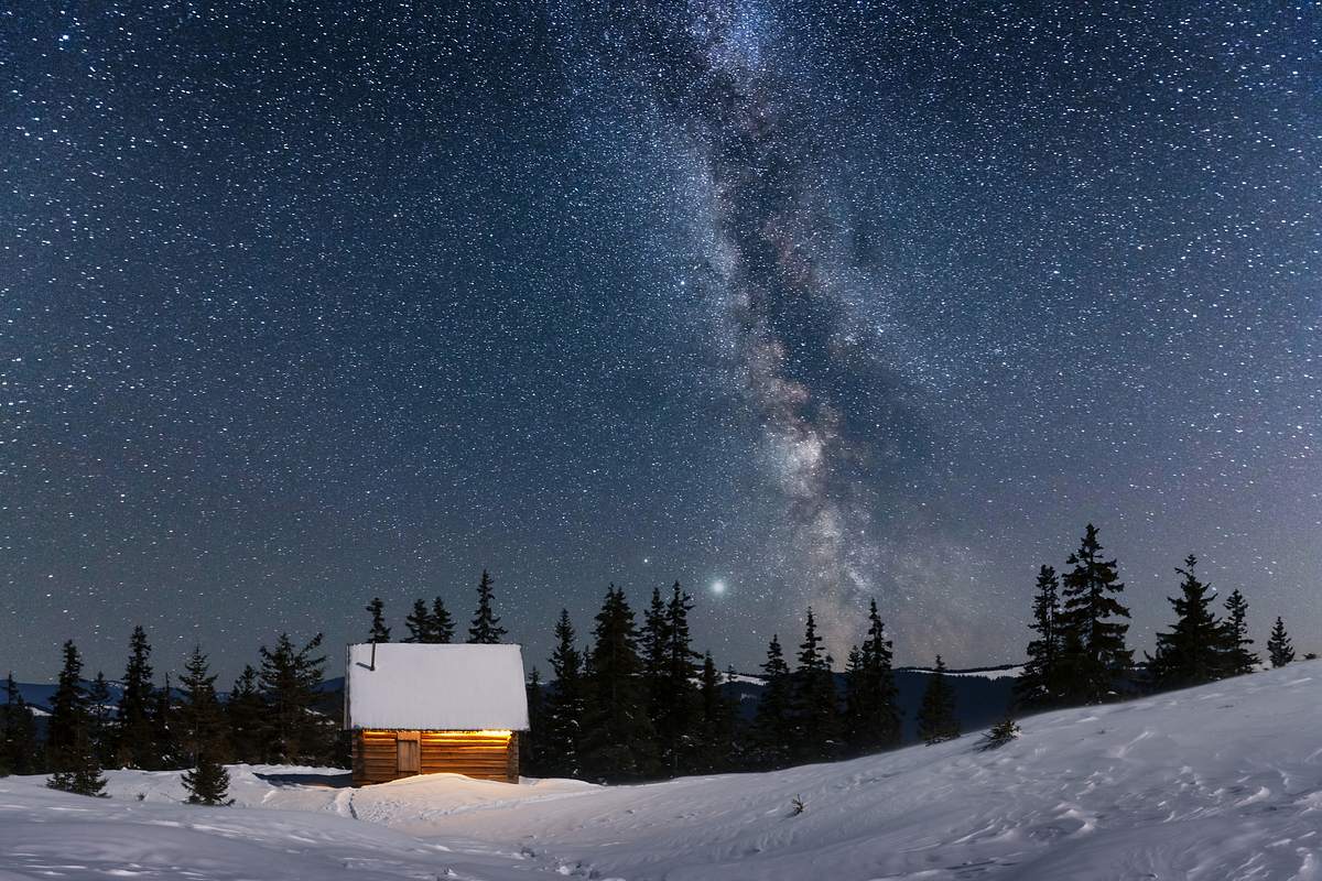 Eine eingeschneite Hütte in einer verschneiten Landschaft unter einem Sternenhimmel.