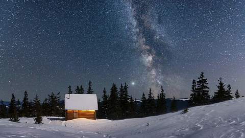 Eine eingeschneite Hütte in einer verschneiten Landschaft unter einem Sternenhimmel. - Foto: iStock / Smitt