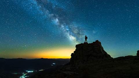 Umriss einer Gestalt auf auf einem Fels blickt auf einen hellen Sternenhimmel - Foto: bjdlzx / iStock