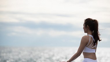 Frau sitzt in Yogapose, den Blick aufs Wasser gerichtet - Foto: sorn340/iStock