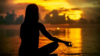 Eine Person meditiert im Schneidersitz am Ufer und genießt die friedliche Stimmung während des Sonnenuntergangs. - Foto: guruXOX / AdobeStock
