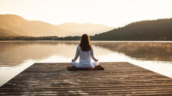 Frau meditiert an einem See - Foto: VisualCommunications/iStock