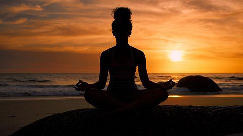 Frau meditiert in Lotuspose am Strand bei Sonnenuntergang - Foto: PeopleImages / iStock