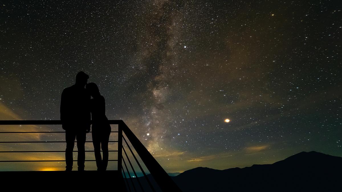 Zwei Personen stehen Arm in Arm auf einer Aussichtsplattorm und blicken auf einen Sternenhimmel.