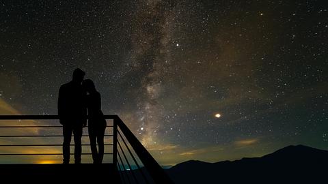 Zwei Personen stehen Arm in Arm auf einer Aussichtsplattorm und blicken auf einen Sternenhimmel. - Foto: Artem Peretiatko/iStock